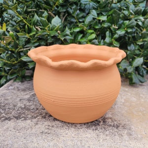 May include: A terracotta flower pot with a scalloped rim and a textured surface. The pot has a light brown color and is made of clay.