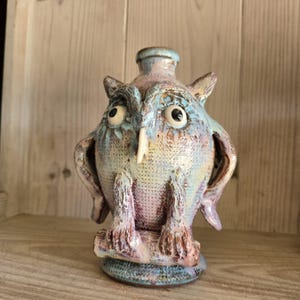 May include: A ceramic owl figurine featuring a textured surface with a mottled glaze in shades of blue, purple, and tan. The owl has large round eyes, a pointed beak, and is perched on a branch-like base with a small opening at the top of its head.