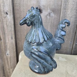 May include: A ceramic rooster figurine in shades of blue and green. The sculpture has a detailed comb, wattle, and ruffled collar. The rooster is perched on a round base, with a glossy finish. The background is a wooden fence.