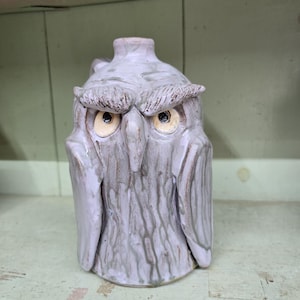 May include: A ceramic jug shaped like an owl with a light lavender glaze and dark brown accents in the textured grooves. The owl features prominent eyebrows, a central beak, and two round eyes with cream-colored irises and black pupils.