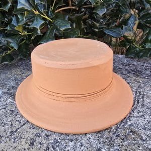 May include: A terracotta-colored, ceramic hat-shaped object with a wide brim and a cylindrical crown. The brim has a series of thin, horizontal lines. The object is sitting on a textured, gray surface, with green foliage in the background.