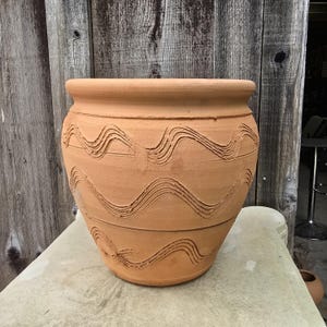 May include: A terracotta-colored, round plant pot with a wide rim and a textured surface. The pot features a series of carved, wavy lines around its body. The pot is sitting on a light-colored surface against a wooden background.