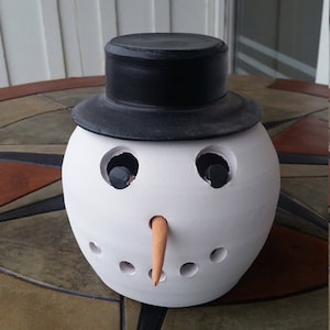 May include: A white ceramic snowman with a black top hat and an orange carrot nose. The snowman has black eyes and a mouth with holes for a candle.