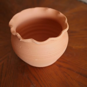May include: A terracotta-colored, round, unglazed ceramic pot with a scalloped rim. The pot has horizontal ridges around its body. This decorative pot is suitable for planting flowers or herbs.