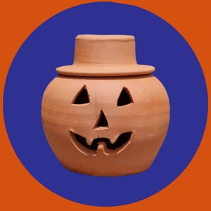 May include: A terracotta pumpkin-shaped candle holder with a carved jack-o'-lantern face and a removable lid. The holder is a light brown color and has a rustic, handmade look.