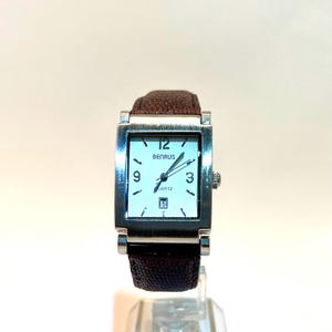 May include: A rectangular Benrus quartz watch with a silver-tone case and a brown leather band. The watch face is white with black numerals and hands. The date is displayed at the 6 o'clock position.