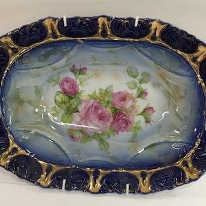 May include: An oval serving dish with two handles. The dish is decorated with a blue and gold rim and a floral design of pink roses on a white background.