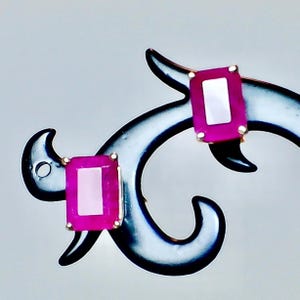 May include: A pair of rectangular, magenta-colored gemstone earrings with a gold-colored setting. The earrings are displayed on a black, decorative stand against a light gray background. The gemstones have a lighter pink center.