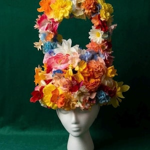 Puede incluir: Un tocado con forma de pesa rusa cubierto por un denso arreglo de flores artificiales en tonos vibrantes de amarillo, naranja, rojo, azul y blanco. La estructura floral presenta un asa central abierta en la parte superior sobre un maniquí blanco.
