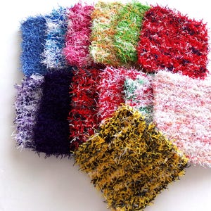 May include: A collection of colorful, square, handmade scrubbies. The scrubbies are in various colors, including blue, pink, green, red, purple, and yellow. Each scrubby has a textured, looped appearance, suggesting they are made from a yarn-like material.