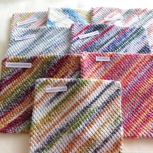 May include: A collection of seven square, hand-crocheted dishcloths featuring diagonal striped patterns in various multi-colored yarn combinations. Each cloth includes a small white label with text such as Sunlit Seafoam, Paris in June, Rosemary Cottage, Moonlit Frost, Walk in the Park, Cinnamon Cedar, Harvest, and Harvest Hues.