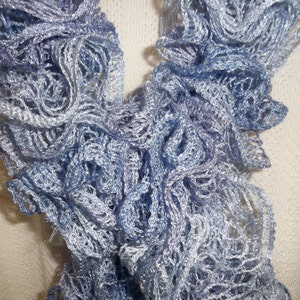 May include: A close-up of a delicate, ruffled scarf in shades of blue and silver. The scarf has a lacy, open weave design, with a textured, layered appearance. The soft, shimmering material suggests a lightweight, elegant accessory.