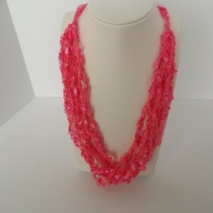 May include: A pink crocheted necklace with a delicate, lacy design. The necklace is made of multiple strands of yarn, creating a layered effect.