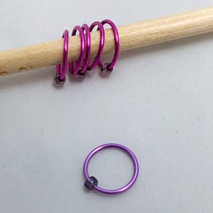 Peut inclure: Un ensemble de cinq marqueurs d'anneaux en métal rose et violet pour le tricot ou le crochet. Les anneaux sont empilés sur une aiguille à tricoter en bois.