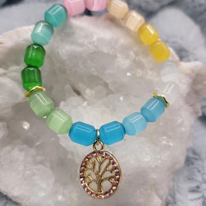 May include: A colorful beaded bracelet with a tree of life charm. The bracelet features a rainbow of colors, including green, blue, pink, and yellow beads. The gold-colored charm is a tree design with pink gemstones.