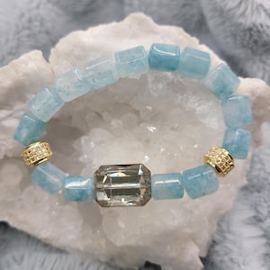 May include: A bracelet featuring light blue, rectangular beads and a large, rectangular, smoky gray crystal. The bracelet has gold-colored spacers with small, sparkling details. The jewelry is displayed on a white, textured surface.
