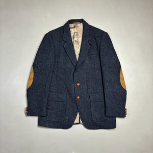 May include: A dark navy blue tweed blazer featuring a textured herringbone pattern, tan suede elbow patches, two orange buttons on the front, two front flap pockets, and a light beige interior lining with a visible brand label at the neck.