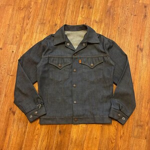 Peut inclure: Une veste en jean gris anthracite avec un col pointu, des manches longues à poignets boutonnés et une fermeture à boutons sur le devant. La veste présente deux poches poitrine à rabats pointus et une petite étiquette orange sur la couture.