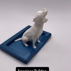 Puede incluir: Una figura geom&eacute;trica blanca de un Bulldog Americano se encuentra sobre una base rectangular azul marino. El perro est&aacute; posando con la cabeza levantada y la boca abierta. La base tiene un borde elevado. El texto "American Bulldog" est&aacute; en un rect&aacute;ngulo negro.