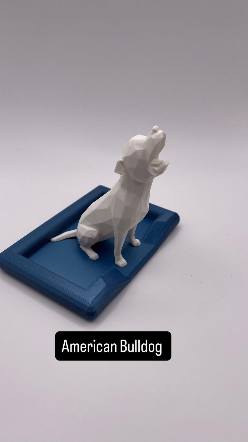 Puede incluir: Una figura geom&eacute;trica blanca de un Bulldog Americano se encuentra sobre una base rectangular azul marino. El perro est&aacute; posando con la cabeza levantada y la boca abierta. La base tiene un borde elevado. El texto "American Bulldog" est&aacute; en un rect&aacute;ngulo negro.