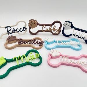 May include: A collection of six bone-shaped name tags in various colors including white, brown, navy, tan, light blue, green, and pink. Each tag features a raised paw print and a custom name in cursive script: Rocco, Cubby, Wilson, Bentley, Dunkin, Miko, and Macarena.