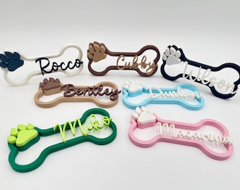 Puede incluir: Una colección de seis etiquetas con forma de hueso en varios colores, incluyendo blanco, marrón, azul marino, tostado, azul claro, verde y rosa. Cada etiqueta presenta una huella de pata en relieve y un nombre personalizado en letra cursiva: Rocco, Cubby, Wilson, Bentley, Dunkin, Miko y Macarena.
