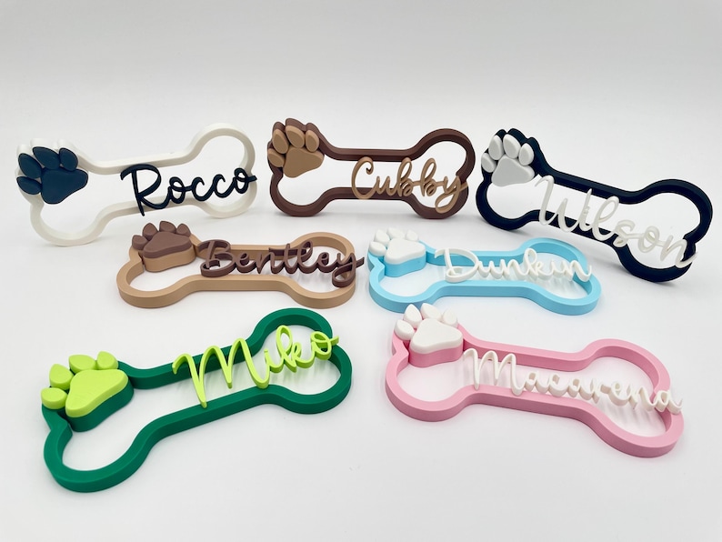 May include: A collection of six bone-shaped name tags in various colors including white, brown, navy, tan, light blue, green, and pink. Each tag features a raised paw print and a custom name in cursive script: Rocco, Cubby, Wilson, Bentley, Dunkin, Miko, and Macarena.
