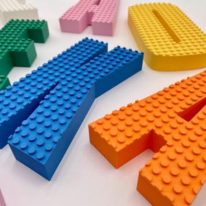 以下が含まれることがあります： 青、オレンジ、黄色、緑、ピンク、白など、さまざまな色の積み木のような形をした立体的な文字のコレクション。各文字の上面はテクスチャ加工されており、小さな円形のスタッドが格子状に並んでいます。
