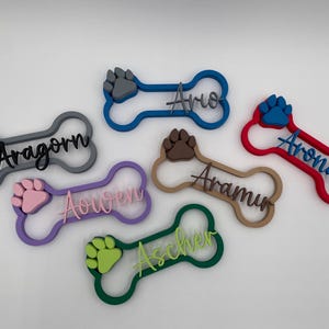 May include: Six bone-shaped pet name tags in various colors, each featuring a raised paw print and a personalized name in script lettering. The tags are colored grey, blue, red, purple, brown, and green with matching or contrasting paw prints.