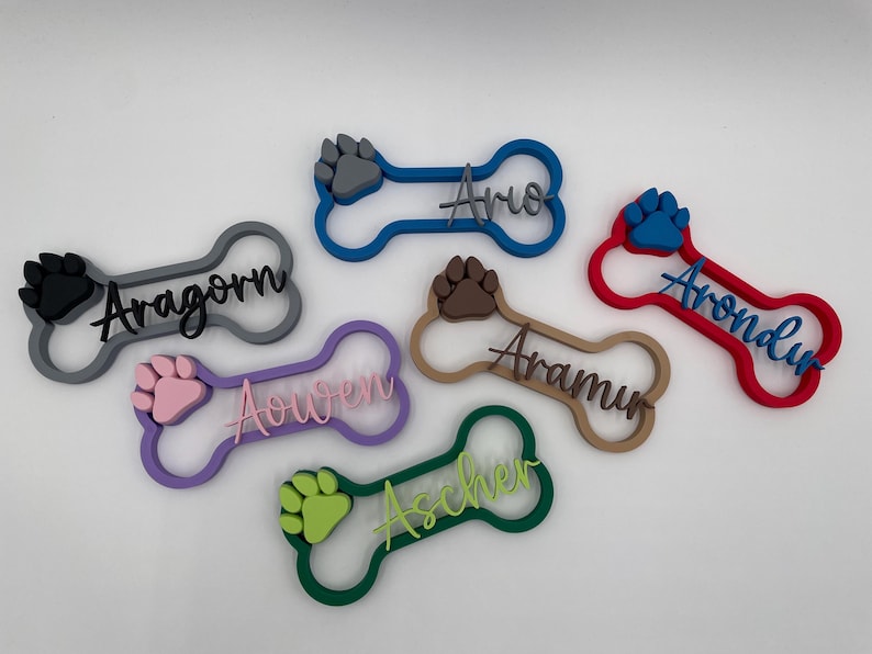 May include: Six bone-shaped pet name tags in various colors, each featuring a raised paw print and a personalized name in script lettering. The tags are colored grey, blue, red, purple, brown, and green with matching or contrasting paw prints.