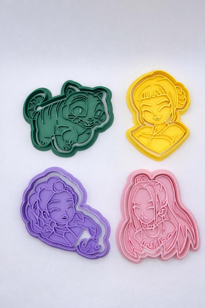 May include: A set of four plastic cookie cutters in various colors. The collection includes a dark green tiger-like creature, a yellow character with a bun hairstyle, a purple character with curly hair, and a pink character with long hair.