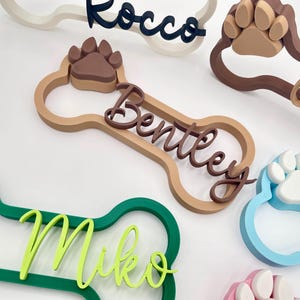 May include: A collection of 3D-printed dog bone-shaped name plaques in various colors. Each plaque features a raised paw print and a custom name in cursive script, including the names Rocco, Bentley, Miko, and others in shades of green, blue, and brown.
