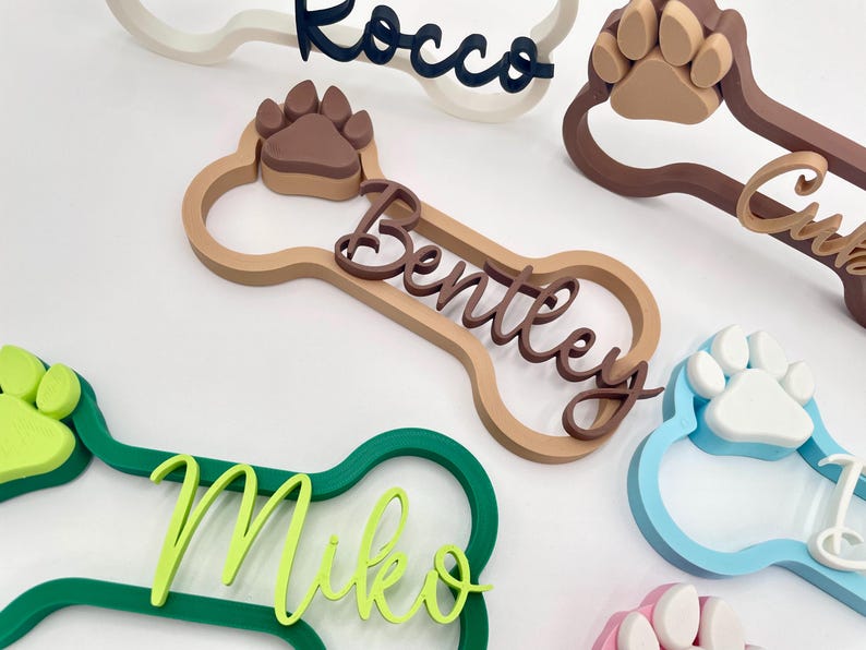 May include: A collection of 3D-printed dog bone-shaped name plaques in various colors. Each plaque features a raised paw print and a custom name in cursive script, including the names Rocco, Bentley, Miko, and others in shades of green, blue, and brown.