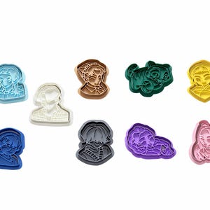 May include: A collection of nine plastic cookie cutters in various colors including light blue, white, brown, dark green, yellow, dark blue, gray, purple, and pink. Each cutter features a unique embossed character design with distinct facial features.