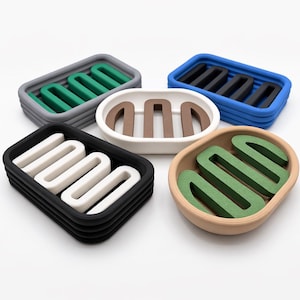 May include: A collection of five oval soap dishes in various color combinations. Each dish features a two-tone design with a solid outer frame and a contrasting wavy, serpentine-shaped insert for drainage, presented in gray, blue, white, black, and tan.