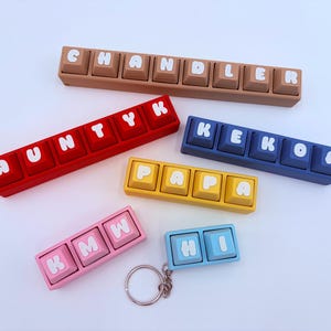 May include: A collection of colorful keyboard keychains spelling out names and words. The keychains are in various colors, including brown, red, yellow, pink, and blue. Each keychain has white letters on the keys. A silver keyring is attached to one of the keychains.