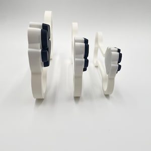 May include: Three white and black paw print shaped cookie cutters arranged in a row from largest to smallest. Each cutter features a white outer frame with a black interior paw pad design, standing upright on their edges to show their profile.