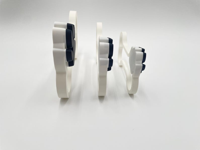 May include: Three white and black paw print shaped cookie cutters arranged in a row from largest to smallest. Each cutter features a white outer frame with a black interior paw pad design, standing upright on their edges to show their profile.