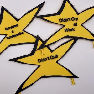 May include: Three yellow star-shaped badges with black outlines and black lettering. The text reads Survived a tummyache, Didn't Cry at Work, and Didn't Quilt. Each star features a small yellow clip attached to one of the points for fastening.