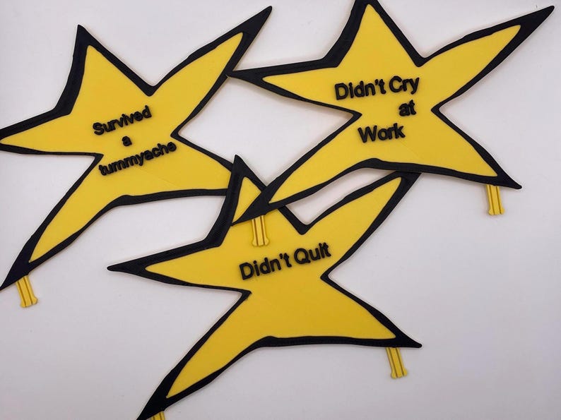 May include: Three yellow star-shaped badges with black outlines and black lettering. The text reads Survived a tummyache, Didn't Cry at Work, and Didn't Quilt. Each star features a small yellow clip attached to one of the points for fastening.