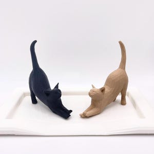 Pu&ograve; includere: Due statuine di gatto in legno in una posa allungata. Un gatto &egrave; nero e l'altro &egrave; di colore marrone chiaro. I gatti sono su un vassoio bianco.