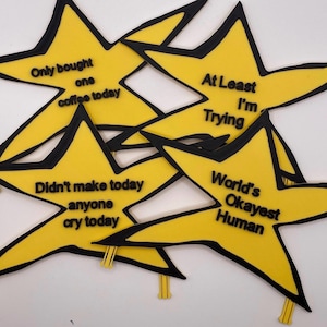 May include: Four yellow star-shaped badges with black outlines and black text. The text reads: Only bought one coffee today, At Least I'm Trying, Didn't make anyone cry today, and World's Okayest Human. Each star has a small yellow clip at the bottom.