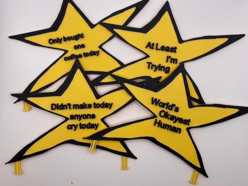 May include: Four yellow star-shaped badges with black outlines and black text. The text reads: Only bought one coffee today, At Least I'm Trying, Didn't make anyone cry today, and World's Okayest Human. Each star has a small yellow clip at the bottom.