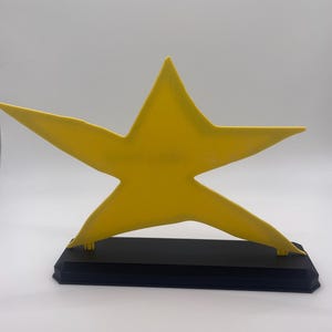May include: A bright yellow five-pointed star figure mounted on a solid black rectangular base. The star has a smooth, matte finish and is positioned upright, centered on the base, creating a clear contrast between the yellow and black colors.