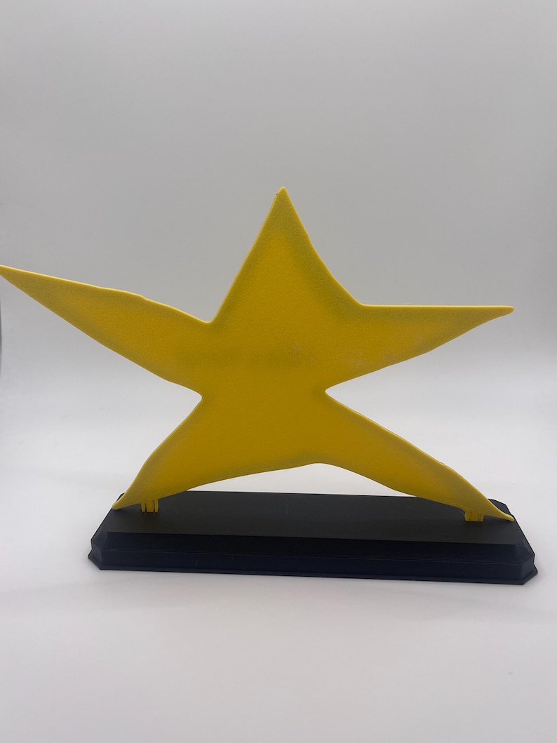 May include: A bright yellow five-pointed star figure mounted on a solid black rectangular base. The star has a smooth, matte finish and is positioned upright, centered on the base, creating a clear contrast between the yellow and black colors.