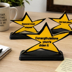 May include: Three yellow star-shaped desk awards with black outlines and black bases. The front star reads Conargthulations youv'e diden it. The back left star reads Second most okayest. The back right star reads Not last place.