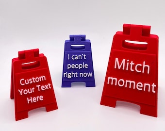 May include: Three miniature A-frame floor signs in red and purple plastic. The left red sign reads Custom Your Text Here, the center purple sign reads I can't people right now, and the right red sign reads Mitch moment in raised white lettering.