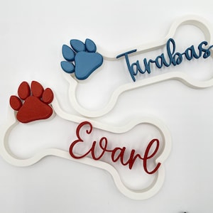 May include: Two white bone-shaped wall signs featuring raised paw prints and personalized names. One sign displays a blue paw print and the name Tarabas in blue script. The other sign features a red paw print and the name Evarl in red script.
