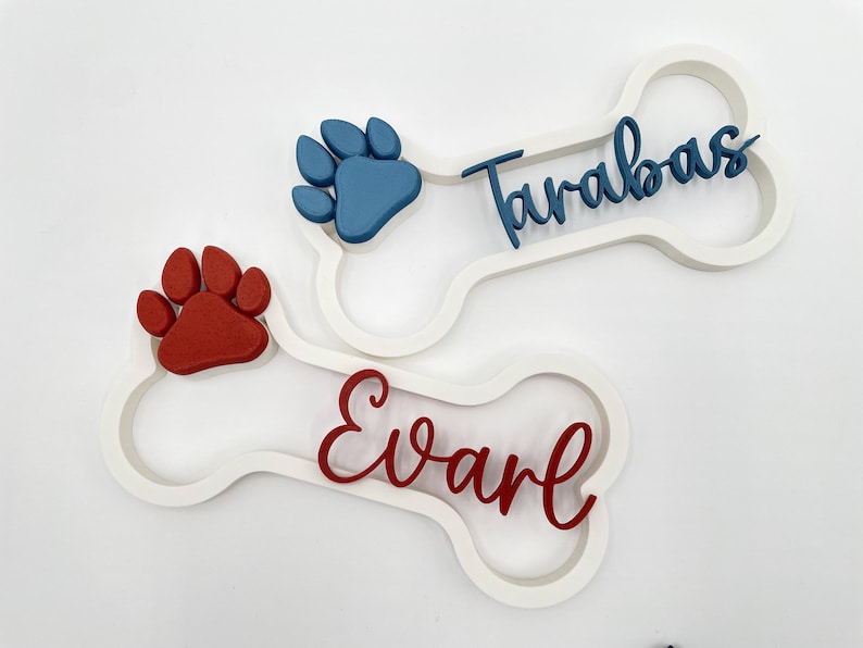 May include: Two white bone-shaped wall signs featuring raised paw prints and personalized names. One sign displays a blue paw print and the name Tarabas in blue script. The other sign features a red paw print and the name Evarl in red script.