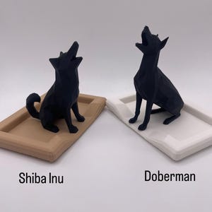 Puede incluir: Dos esculturas geom&eacute;tricas negras de perros, un Shiba Inu sobre una bandeja beige y un Doberman sobre una bandeja blanca. Ambos perros est&aacute;n en posici&oacute;n de aullido. Los textos "Shiba Inu" y "Doberman" est&aacute;n debajo.